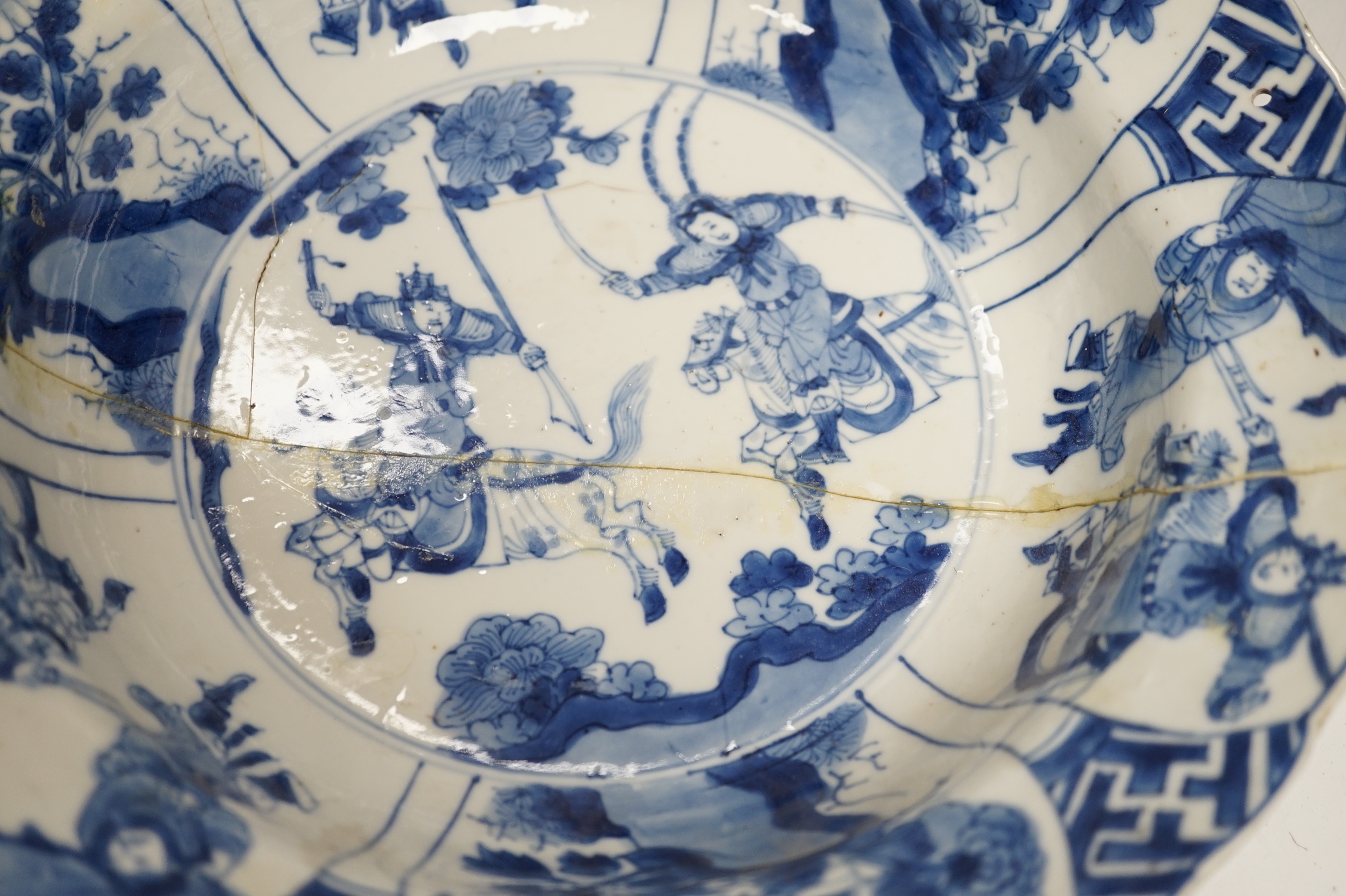 A Chinese blue and white ‘warriors’ bowl, Kangxi mark and period, a similar ‘hunting’ saucer dish, and a famille verte saucer dish, all Kangxi period (3), largest 27cm in diameter